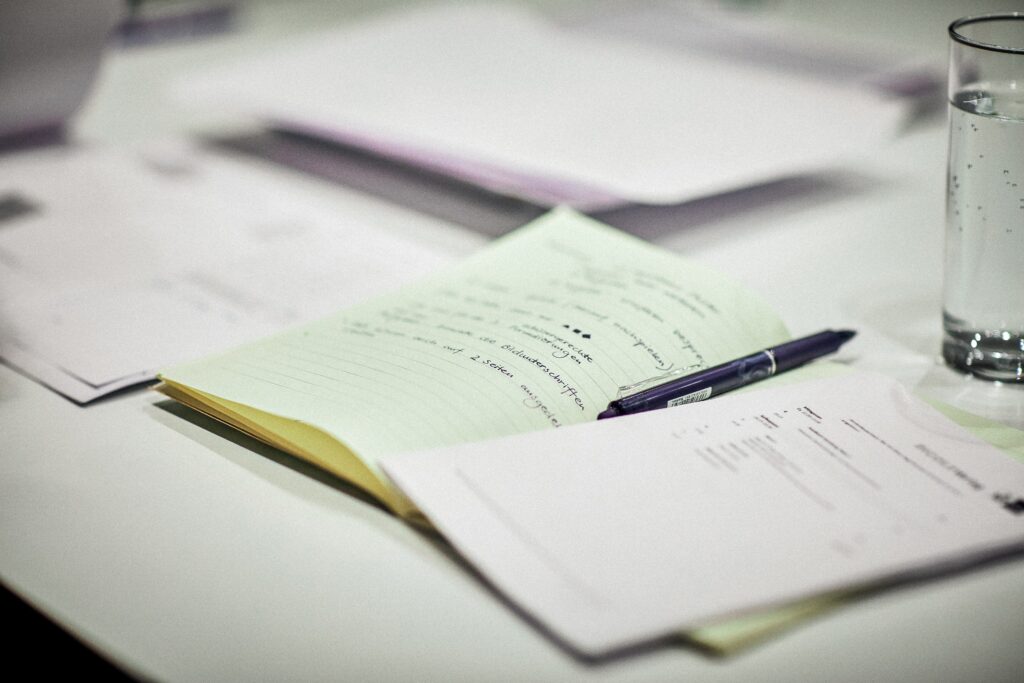 Notebook with notes on a white table. Blue pen rests on notebook. Papers and a glass of water on the background.