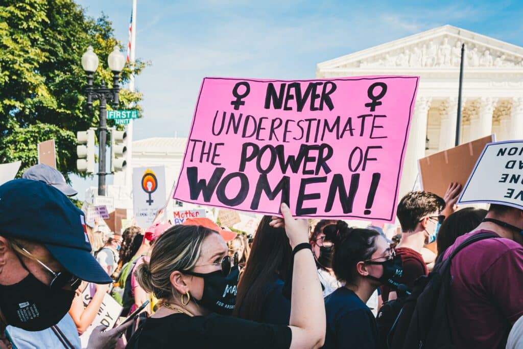 A protest scene outside a government building, with a crowd of people wearing masks. A woman in the foreground holds up a pink sign that reads 'NEVER UNDERESTIMATE THE POWER OF WOMEN!' with feminist symbols on either side.