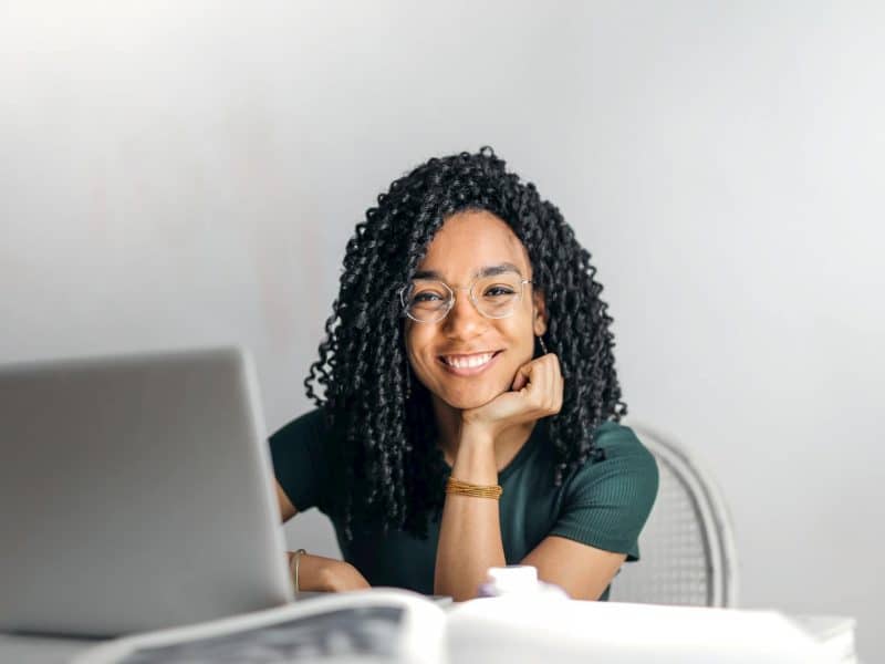 A young Black woman smiling and sitting at a desk with a laptop. Caption: Photo by Andrea Piacquadio on Pexels.