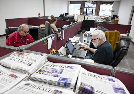 People working at desks in a newspaper office with printed newspapers in the foreground.