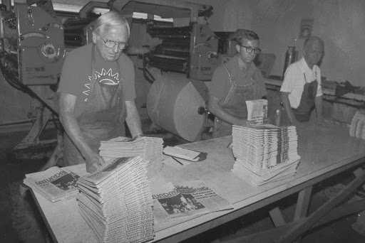 Three men stand in front of a printing press, assembling newspaper stacks on a table.